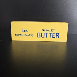 May include: A rectangular yellow block resembling a stick of butter with blue text that reads 4 oz. Net Wt. (One Elf) Salted Elf BUTTER. The object has a smooth, solid appearance and is presented against a dark, neutral background.