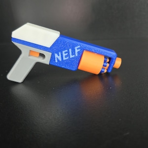 May include: A toy blaster featuring a blue body with the white text NELF printed on the side, a white top section, a gray handle, and an orange rotating cylinder containing small blue foam darts with orange tips.