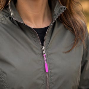 May include: An olive green lightweight jacket featuring a high collar and a full-length front zip. The zip pull is accented with a bright neon pink rectangular tab attached by a thin matching cord, contrasting against the muted fabric.