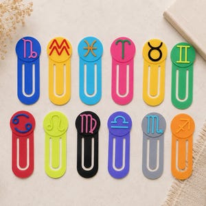May include: A collection of colorful zodiac sign bookmarks. Each bookmark features a different sign symbol in a contrasting color, such as blue, yellow, pink, and green. The bookmarks are arranged on a light-colored surface, with a book and decorative elements in the background.