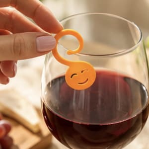 May include: An orange plastic wine glass charm featuring a circular face with a smiling expression and a curved hook attachment. The charm is designed to hang from the rim of a glass filled with red wine, serving as a marker for identification.