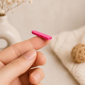 May include: A small, bright pink plastic lancet device, held between fingers. The lancet is rectangular with two small holes on one end and a single hole on the other. The background includes a vase with flowers and a ball of twine.