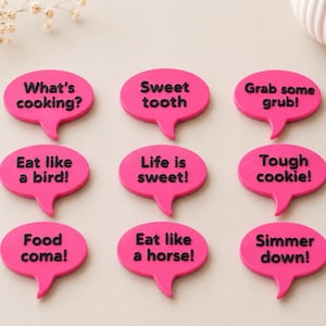 May include: Nine bright pink speech bubble magnets with black text. The phrases include What's cooking?, Sweet tooth, Grab some grub!, Eat like a bird!, Life is sweet!, Tough cookie!, Food coma!, Eat like a horse!, and Simmer down!.