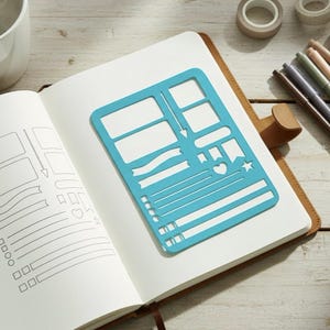 May include: A bright blue plastic stencil featuring various rectangular boxes, banners, checklist lines, a heart, and a star shape, resting on the open page of a notebook where the same shapes are traced in black ink on the left side.