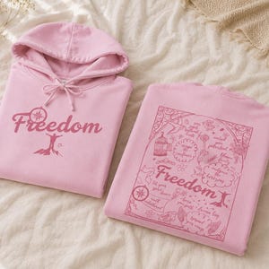 Puede incluir: Una sudadera con capucha rosa claro que presenta un gráfico rosa oscuro en la parte delantera con la palabra Freedom y una persona sobre una montaña. La parte trasera muestra un estampado detallado con la palabra Freedom y frases inspiradoras.