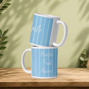 Puede incluir: Dos tazas de cerámica blanca apiladas verticalmente, con un patrón de rayas verticales en azul claro y blanco. La taza superior muestra la palabra Smile en letra cursiva blanca, mientras que la inferior tiene las palabras Breathe, Trust, Advance.