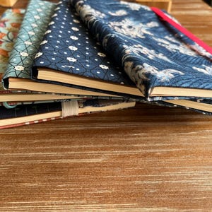 Può includere: Una pila di cinque quaderni con copertine a motivi diversi. Le copertine sono blu, nere e bianche con motivi floreali e geometrici. I quaderni sono impilati su una superficie di legno.