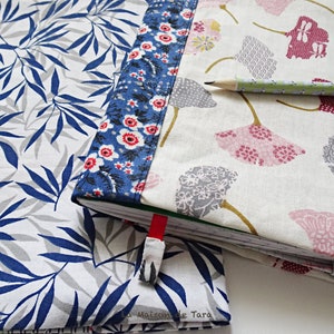 May include: Three fabric-covered notebooks with different patterns. The notebook on the left has a blue and white leaf pattern. The notebook in the middle has a blue and white floral pattern. The notebook on the right has a white background with pink and gray ginkgo leaf patterns. The text "La Maison de Tara" is visible on the bottom left notebook.