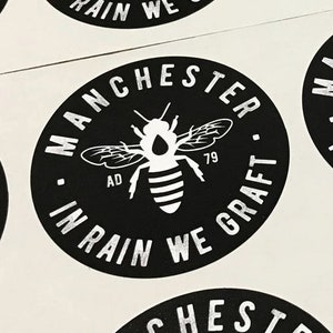 May include: Black and white sticker with a bee and the text "Manchester In Rain We Graft" around the bee.