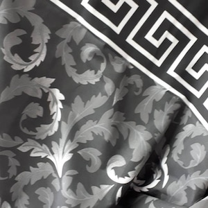 Könnte beinhalten: Dunkelgrauer Stoff mit einem sich wiederholenden silbernen Blumenmuster und einem weißen griechischen Schlüsselbordürenrand. Der Stoff ist gefaltet und zeigt das Muster und die Bordüre im Detail. Der Stoff hat ein luxuriöses Aussehen.