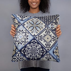 Puede incluir: Cojín decorativo cuadrado con un patrón de azulejos geométricos en tonos azul, crema y marino. El cojín presenta una variedad de diseños florales y de volutas ornamentados dentro de las formas cuadradas y de diamante. El cojín es sostenido por una persona.
