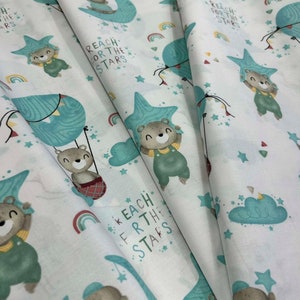 Op de afbeelding: Witte stof met een herhaald patroon van cartoonberen, sterren, regenbogen en wolken. De beren dragen overalls en ster-vormige hoeden. De tekst luidt "REACH FOR THE STARS". De stof heeft een grillig, speels ontwerp.