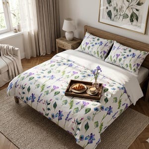May include: A double bed dressed with a floral duvet cover and matching pillowcases. The white bedding is decorated with watercolour-style purple, blue, and green botanical patterns. A wooden tray with a croissant and coffee sits on the bed, creating a cosy atmosphere.