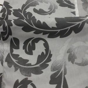 Könnte beinhalten: Nahaufnahme eines weißen Stoffes mit einem sich wiederholenden Muster aus grauen, stilisierten Blättern und Ranken. Die Blätter haben ein strukturiertes Aussehen, und das Design ist auf den Stoff gedruckt. Der Stoff ist gefaltet und zeigt die Wiederholung des Musters.