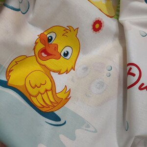 May include: White fabric featuring a cartoon duck illustration. The duck is yellow with an orange beak and blue eyes, set against a blue and white water background. Other elements include a red flower and the word "Duck".