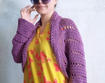 May include: A purple crochet shrug with long sleeves. The shrug is worn over a yellow dress with a red butterfly pattern. The person is wearing large black sunglasses.