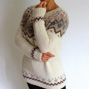 Wrap yourself in softness with this stunning hand-knit Icelandic-style sweater in creamy white tones with subtle Fair Isle patterns in earthy brown, soft grey, and blush pink. This cozy pullover is the perfect blend of rustic charm and modern elegance – a beautiful staple for winter.
