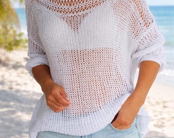 May include: A white, open-weave knit sweater with a round neckline and long sleeves. The sweater is paired with light blue denim shorts. The beach setting includes palm trees and the ocean in the background.
