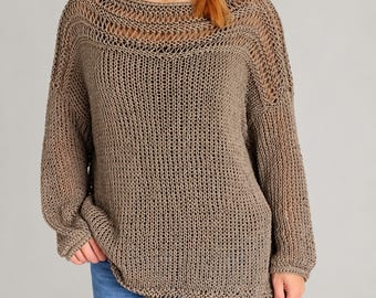May include: A taupe-colored, long-sleeved sweater featuring an open-knit, loose weave construction. The garment has a wide, rounded neckline with a decorative horizontal stitch pattern across the upper chest and shoulders, paired with blue jeans.