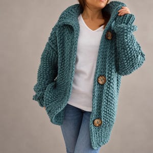 May include: A chunky knit teal cardigan featuring a wide collar, long sleeves, and three large, round, mottled brown buttons down the front. The garment is worn over a plain white v-neck t-shirt and light blue denim jeans, showcasing a textured stitch.
