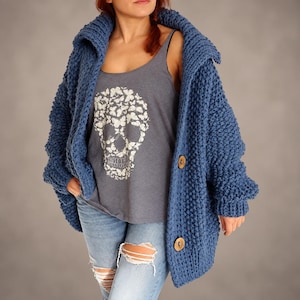 May include: A chunky knit blue cardigan with a wide collar and two large wooden buttons. Underneath, a grey tank top features a white graphic print of a skull composed of small butterflies. Light blue distressed denim jeans with knee rips.