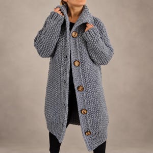Könnte beinhalten: Eine langärmelige, grob gestrickte Strickjacke in staubigem Blau. Das Kleidungsstück verfügt über einen markanten Kragen, ein strukturiertes Noppenmuster und fünf große, runde, braune Holzknöpfe an der Vorderseite über einer schwarzen Basis.