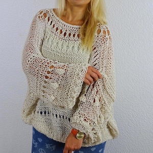 Woman wearing an openwork knit sweater with her arms crossed, showing its flowing sleeves and decorative knit bands, paired with patterned blue trousers.