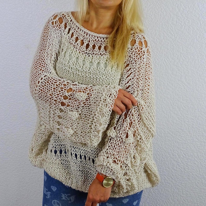 Woman wearing an openwork knit sweater with her arms crossed, showing its flowing sleeves and decorative knit bands, paired with patterned blue trousers.