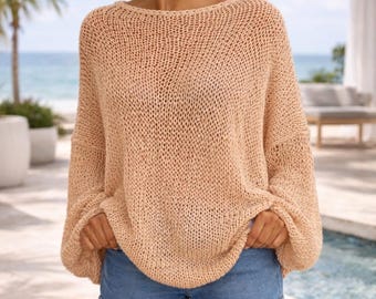 May include: A light peach-colored knit sweater with a wide neckline and long sleeves. The sweater is paired with denim shorts. The image is taken outdoors with a beach and ocean in the background.