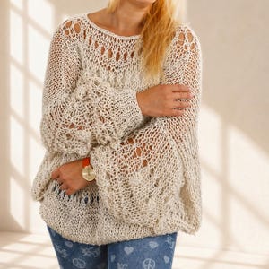Hand knit off white cotton boho sweater with oversized fit, open knit texture and statement bell sleeves, shown with crossed arms in soft natural window light.