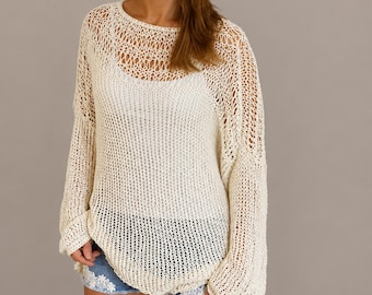 May include: An off-white, long-sleeved, loose-knit sweater featuring a wide, open-weave pattern. The sweater has a rounded neckline and is worn over a solid white camisole. The garment is paired with light blue denim shorts with white lace trim.