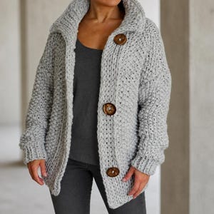May include: A light gray chunky knit cardigan featuring a wide collar and three large, round, dark brown wooden buttons down the front. The garment is crafted with a thick, textured yarn and has long sleeves with ribbed cuffs, worn over a dark gray top.