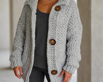 May include: A light gray chunky knit cardigan featuring a wide collar and three large, round, dark brown wooden buttons down the front. The garment is crafted with a thick, textured yarn and has long sleeves with ribbed cuffs, worn over a dark gray top.