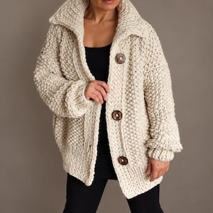 May include: A chunky knit cream-colored cardigan featuring a wide collar, long sleeves, and three large, round, dark brown buttons down the front. The garment is crafted with a textured, thick yarn stitch pattern and is worn over a black top.