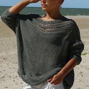 Könnte beinhalten: Ein locker sitzender, olivgrüner Strickpullover mit rundem Ausschnitt und langen Ärmeln. Der Pullover hat ein strukturiertes Strickmuster und eine entspannte Passform. Der Halsausschnitt hat ein dekoratives offenes Strickdesign. Der Pullover wird am Strand getragen.