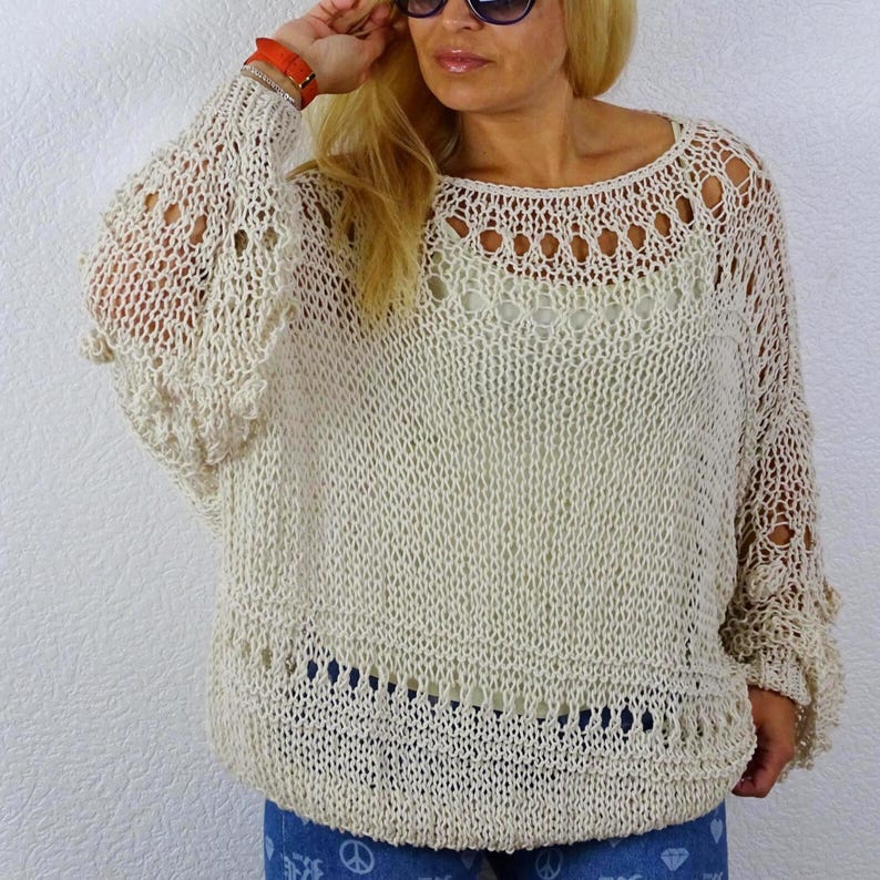 Front view of a woman wearing an openwork knit sweater with a wide neckline, decorative knit bands and loose sleeves, paired with patterned blue trousers.