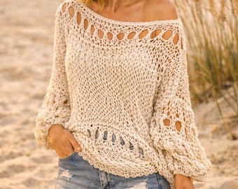 May include: Off-white, open-knit sweater with a boat neck and long, bell sleeves. The sweater has a textured, woven appearance with decorative cut-out details along the neckline and hem. The model is wearing denim shorts.