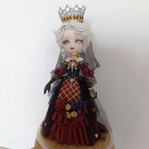 May include: A decorative doll with white hair, a silver crown, and a grey veil. The doll wears a dark red and black gown with a gold lattice pattern, ruffled layers, and fabric roses. The bodice features intricate gold and red embellishments.