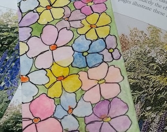 May include: A bookmark with a green background and a colorful floral pattern. The flowers are painted in shades of pink, yellow, blue, purple, and white. The bookmark has a yellow string attached to it.