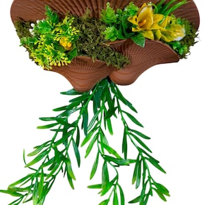 May include: A brown, 3D-printed wall planter shaped like a mushroom cap, featuring a textured, ribbed surface. The planter is filled with artificial green moss, small yellow flowers, and vibrant green leafy sprigs that hang downward.