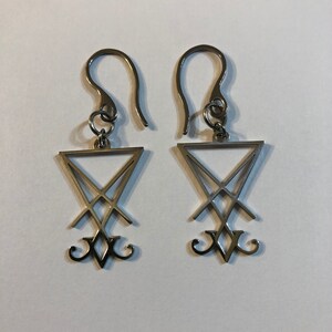 Puede incluir: Un par de pendientes de plata con un colgante triangular que presenta un dise&ntilde;o de pentagrama invertido estilizado.