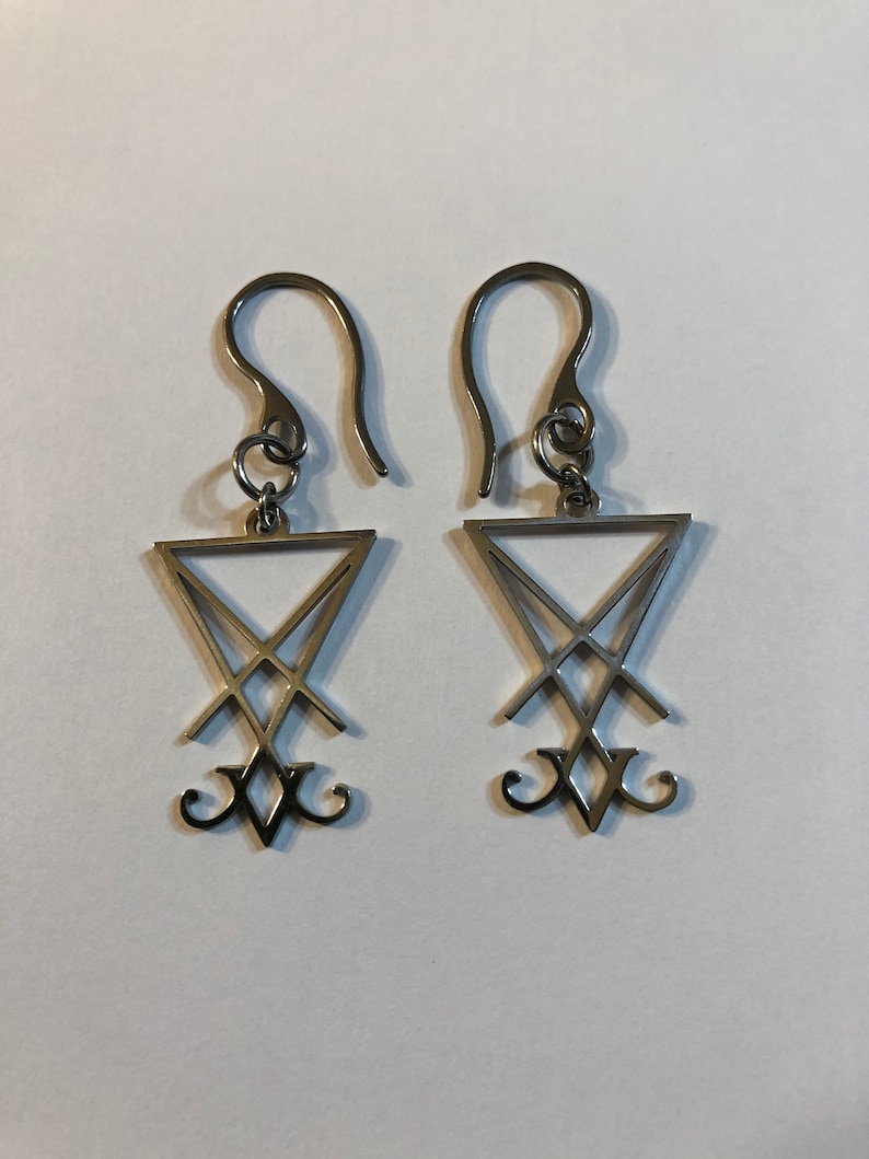 Puede incluir: Un par de pendientes de plata con un colgante triangular que presenta un dise&ntilde;o de pentagrama invertido estilizado.