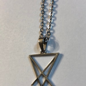 Puede incluir: Un collar de cadena de plata con un colgante de plata que presenta un s&iacute;mbolo de pentagrama con una 'V' estilizada en la parte inferior.
