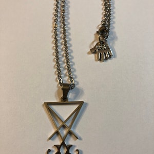 Puede incluir: Un collar de plata con un colgante que presenta el sello del demonio Leviat&aacute;n. El colgante es un tri&aacute;ngulo con una cruz en su interior y una 'V' estilizada en la parte inferior. El collar tambi&eacute;n tiene un peque&ntilde;o amuleto que dice 'Hand Made'.