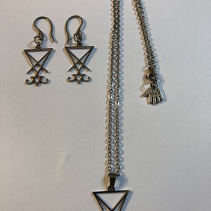 Puede incluir: Un conjunto de collar y pendientes de plata con el sello del demonio Baphomet. El collar tiene una cadena con un peque&ntilde;o amuleto de mano y un colgante con el sello. Los pendientes son simples ganchos con el sello colgando de ellos.