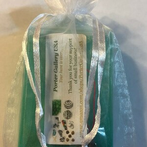 Puede incluir: Una peque&ntilde;a caja verde con una etiqueta blanca que dice "Porter Gallery USA" y "Your hunt is out." La etiqueta tambi&eacute;n incluye un mensaje de agradecimiento por el apoyo a las peque&ntilde;as empresas y una direcci&oacute;n de sitio web. La caja est&aacute; en una bolsa de organza blanca con lazos de cinta blanca.