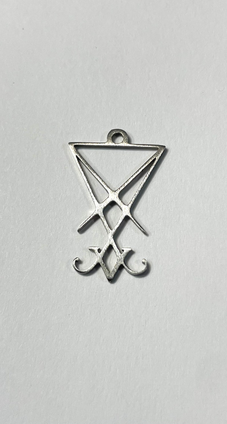 Puede incluir: Un colgante de plata con un pentagrama invertido estilizado con una cruz en su interior. El colgante tiene un bucle en la parte superior para sujetarlo a una cadena.