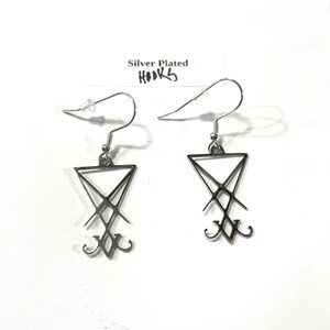 Puede incluir: Un par de pendientes plateados con un dise&ntilde;o de pentagrama. Los pendientes est&aacute;n sobre un fondo blanco con una etiqueta que dice "Silver Plated Hooks".