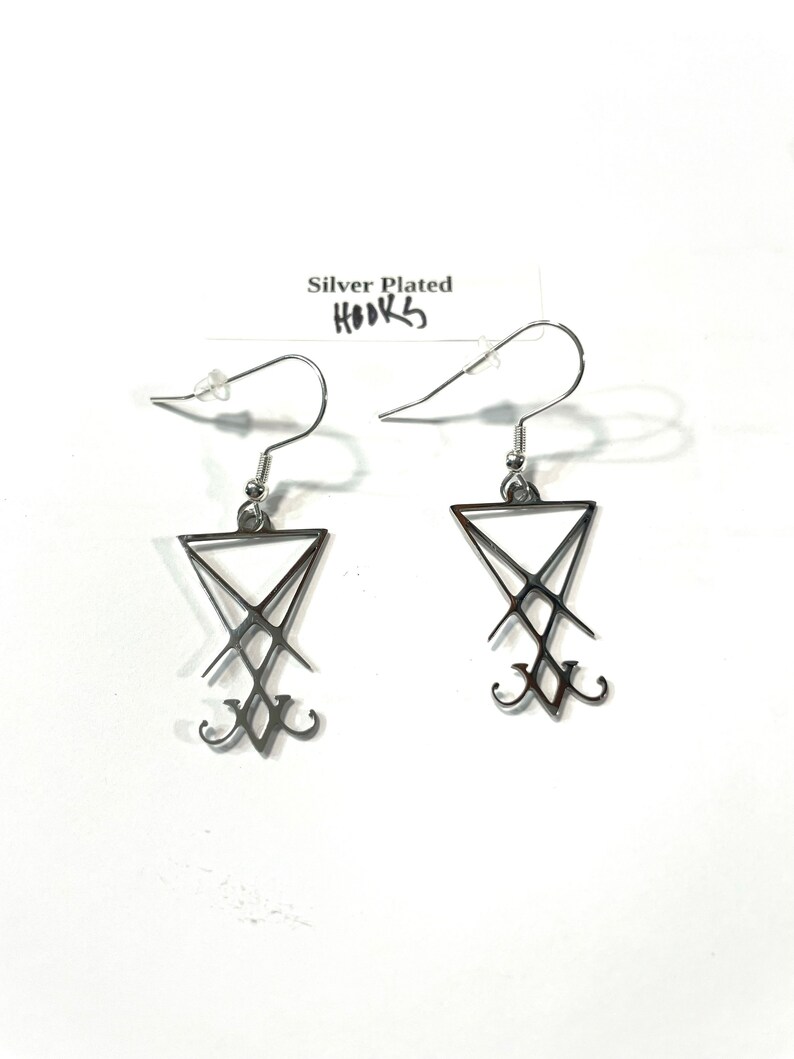 Puede incluir: Un par de pendientes plateados con un dise&ntilde;o de pentagrama. Los pendientes est&aacute;n sobre un fondo blanco con una etiqueta que dice "Silver Plated Hooks".