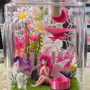 May include: A miniature fairy garden scene inside a clear plastic cube. A pink fairy with white wings sits on green moss with a white rabbit and a purple mushroom. The garden also includes pink flowers, a white picket fence, and a pink bird.